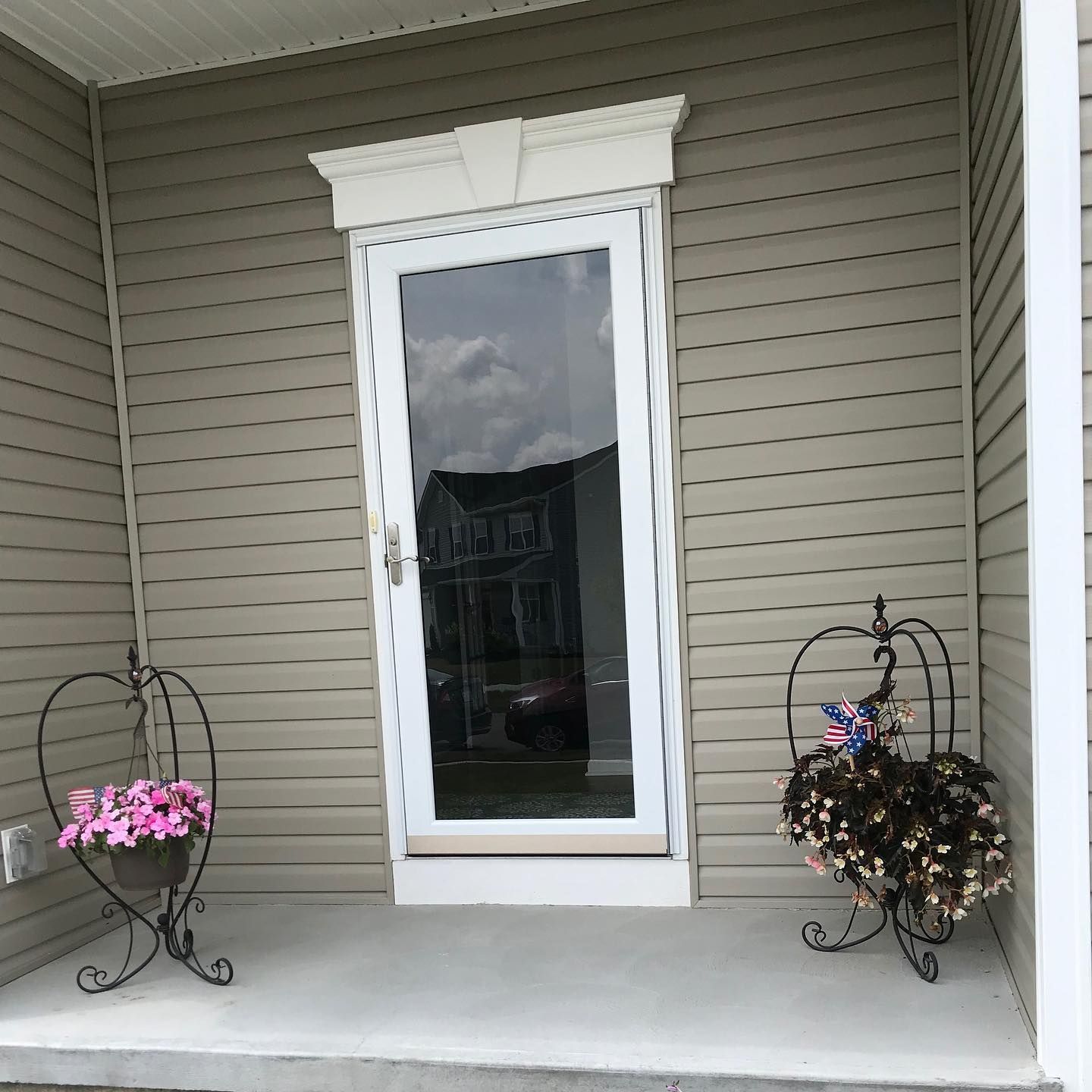 A light brown, vinyl-sided porch entrance with a white storm door, flanked by two decorative iron plant stands with flowers.