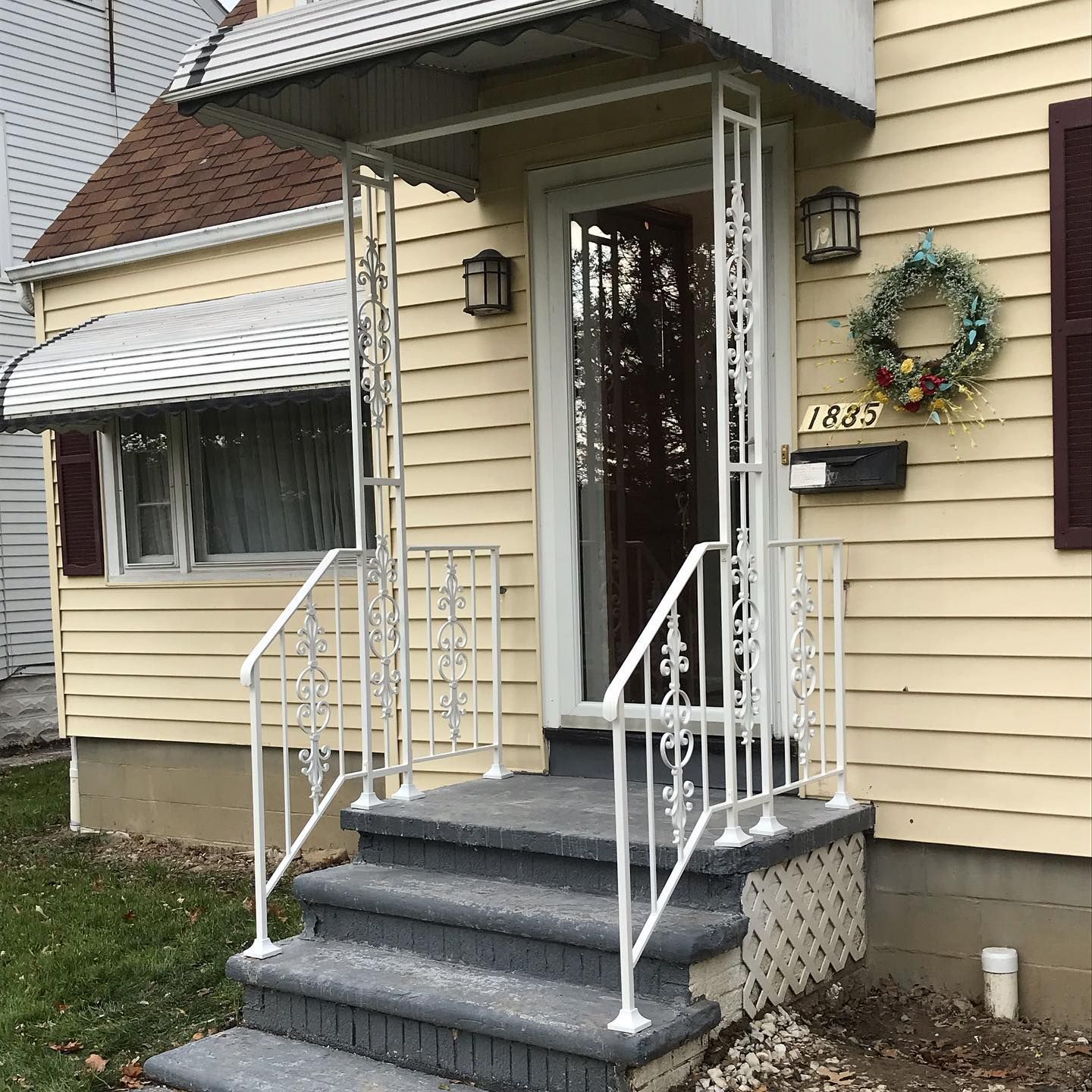 A yellow house with a concrete stoop, white metal railings, a small awning over the front door, and a decorative wreath.