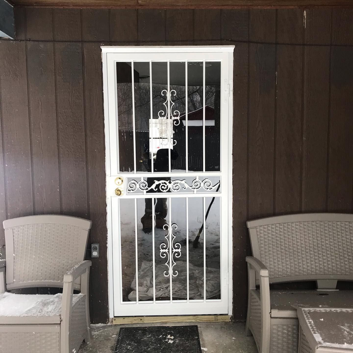 A white metal security storm door with decorative scrollwork sits in a brown wall between two tan patio chairs.