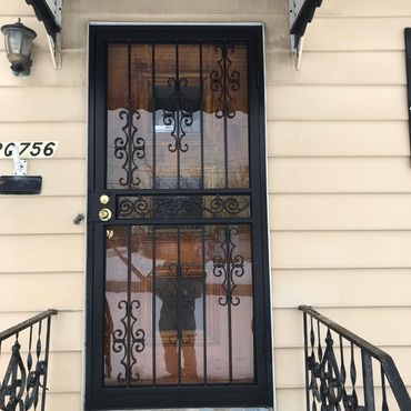 Black front door with decorative iron grille on a beige building entrance, flanked by metal railings. Black front door with decorative iron grille on a beige building entrance, flanked by metal railings.