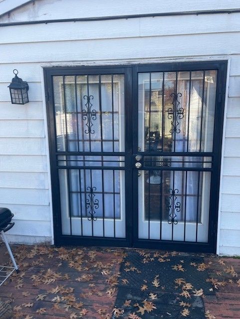 Black metal double front doors with glass panels and a small wall lantern on a white house exterior.