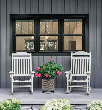Two white rocking chairs on a porch with a potted pink flower arrangement in front of a black window Two white rocking chairs on a porch with a potted pink flower arrangement in front of a black window