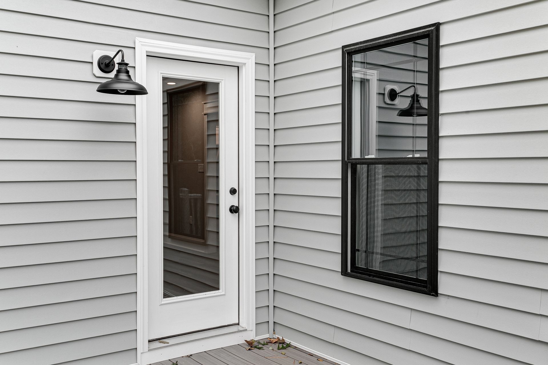 White house entry with glass door, black wall light, and a black-trimmed window on gray siding