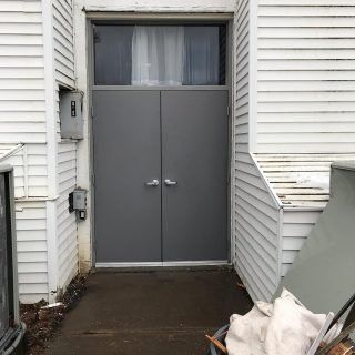 Gray double doors at the entrance of a house, with white siding and a wet walkway.