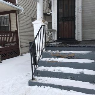 Snow-covered front steps leading to a house entrance with black railings and a white porch column