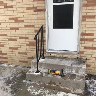 Concrete steps with a black railing leading to a white door on a brick wall; a yellow tool lies on the step.