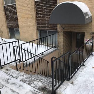 Snowy apartment entrance with black railings, steps, and a dark awning over the doorway