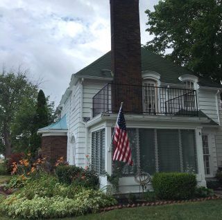 White house with green roof, balcony, American flag, and tall brick chimney amid lush garden greenery