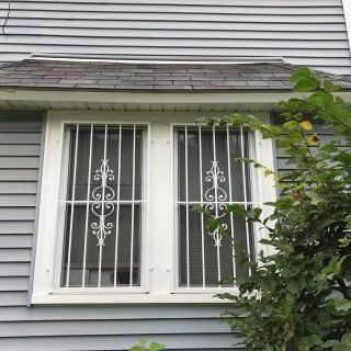 House window with white security bars, gray siding, and a leafy tree partly covering the right side