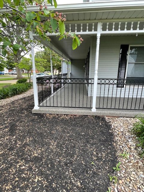 Front porch with black railing, white trim, and a mulched front yard beneath a tree