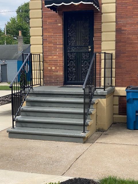 Brick house front entrance with black iron railing, concrete steps, and a blue trash bin by the sidewalk.