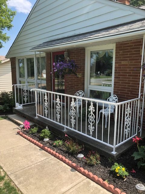 Front porch with white railing, hanging flowers, and a brick facade beside a concrete walkway.