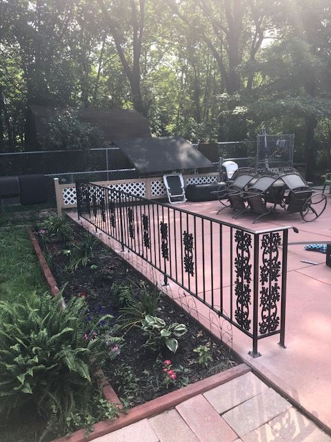 Patio with black metal railing, lounge chairs, and grill surrounded by trees and garden beds