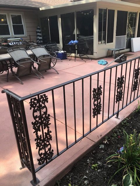 Patio with black metal railing, pink floor, patio chairs, and sliding glass doors beside a garden