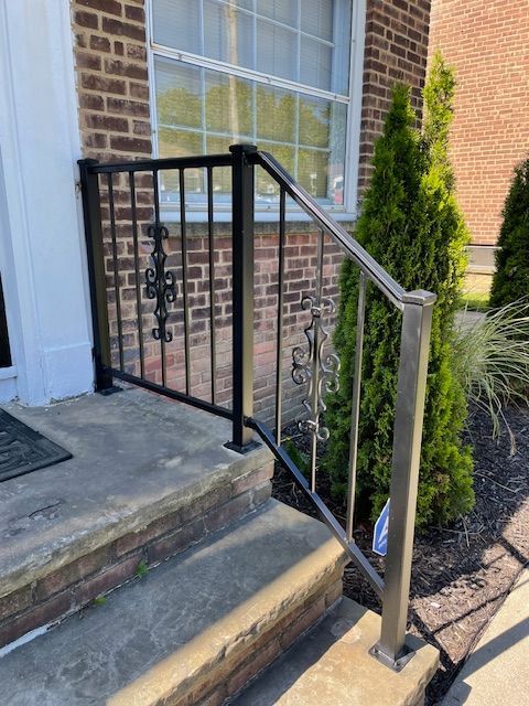 Front porch steps with a black metal handrail and silver support post beside a brick house.