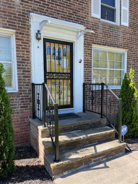 Brick townhouse entrance with black iron gate, steps, and two shrubs.
