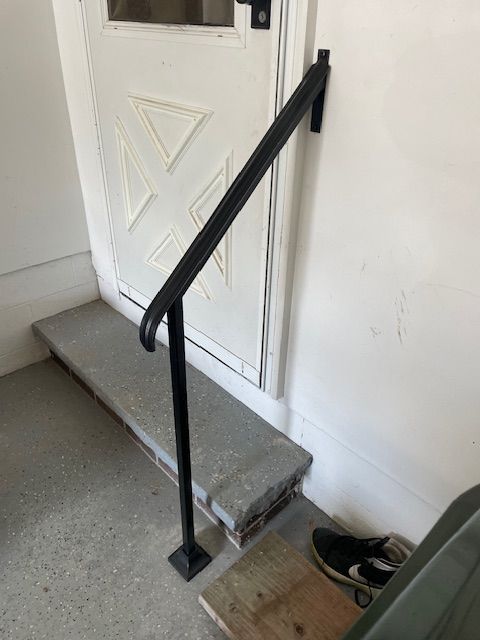 Black handrail beside a small concrete step leading to a white door with shoes nearby