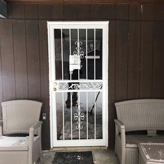 Front porch with a white security door, dark wood siding, and wicker chairs on either side.