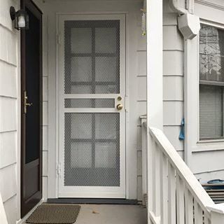 Front porch with white screen door, gray siding, window, and railing beside a dark entry door.