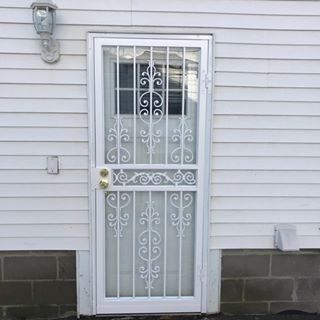 White metal security door with decorative grill on gray siding, with wall light and basement windows beside it