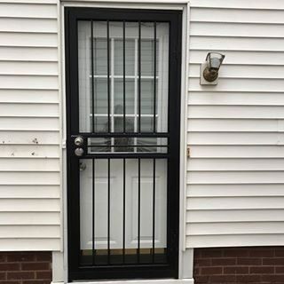 Black front door with security bars beside a wall-mounted outdoor light on a brick and vinyl exterior.
