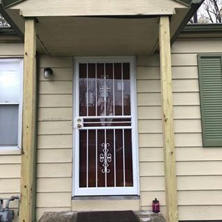 Front porch entrance with white security door, beige siding, and green shutters under a small awning