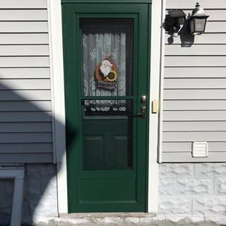 Green front door with a lace-curtained window and wreath on a white house exterior