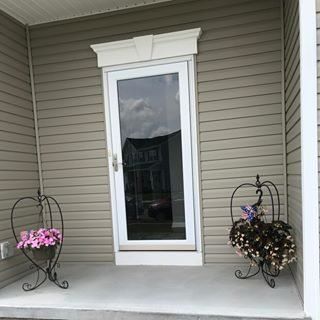 Front porch with a white-framed glass door, beige siding, and two decorative flower stands with potted blooms.