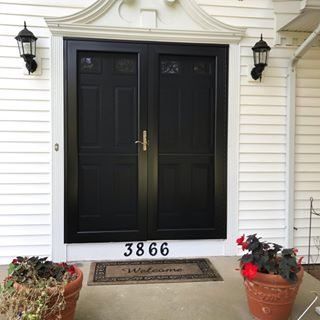 Black double front doors with brass handles on a white house, flanked by lanterns and potted plants.