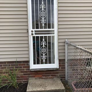 White security door with decorative bars beside a chain-link fence on a brick-and-siding house.