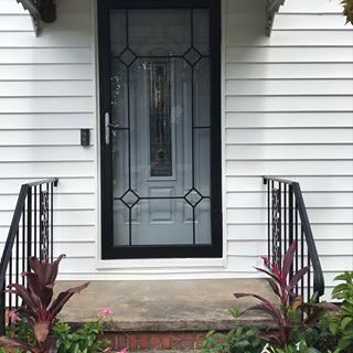 Front porch with black-framed glass door, white siding, and potted plants by the steps.