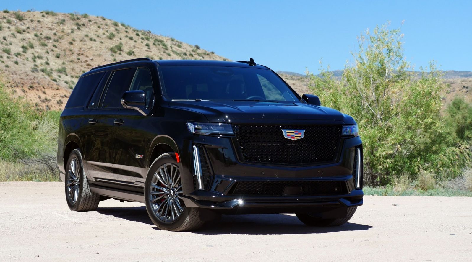 Black Cadillac Escalade SUV parked on a dirt road in a sunny desert setting.
