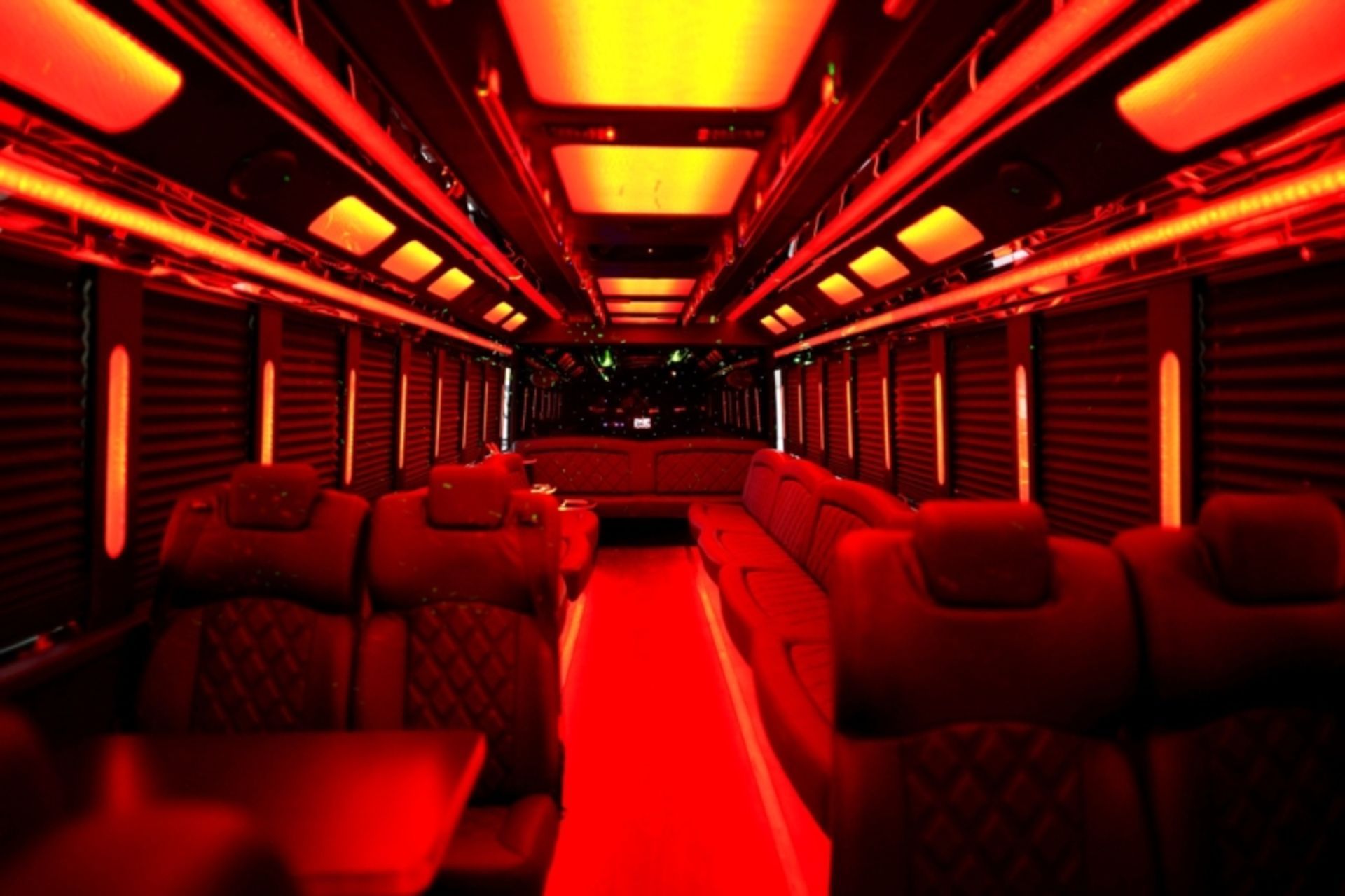 Inside of a red-lit luxury bus with plush, red seating and overhead lighting. A dark hallway leads to the back.