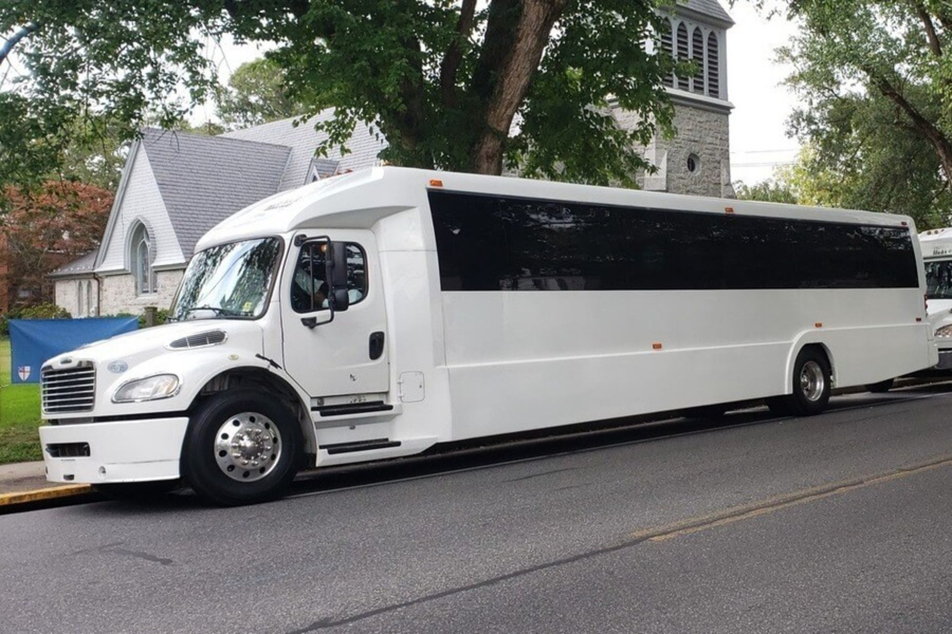 White party bus parked on a street, with a church and trees in the background.