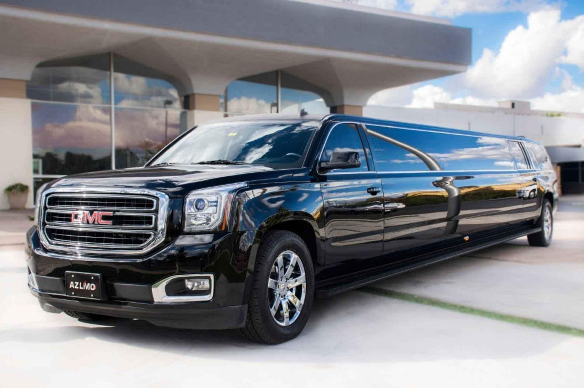 Black GMC Yukon limousine parked in front of a modern building with large windows, under a bright sky.