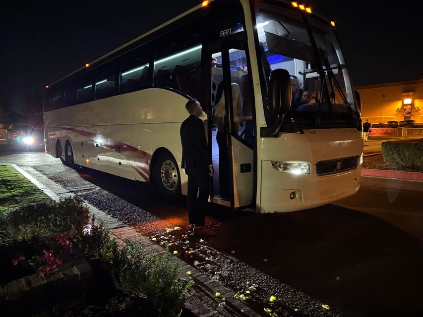 A white tour bus is parked at night with its door open. A person in a dark suit stands in the doorway, looking outward.