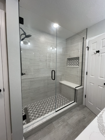 A glass-enclosed shower with light gray tile walls, a patterned tile floor, a built-in bench, and black hardware.