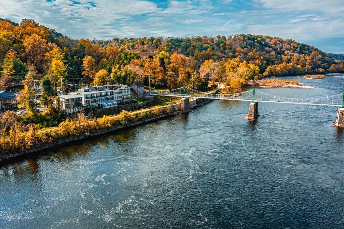 River view with autumn trees, a white building on the left, and bridge piers in the water under a blue sky in Bucks County