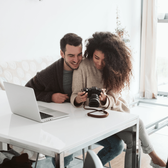 A man and a woman are sitting at a table with a laptop and a camera.