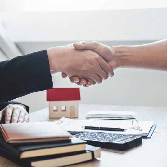 Two people shaking hands over a table with a small house in the background.