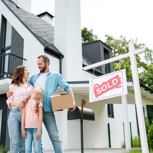 A family is standing in front of a house with a sold sign.