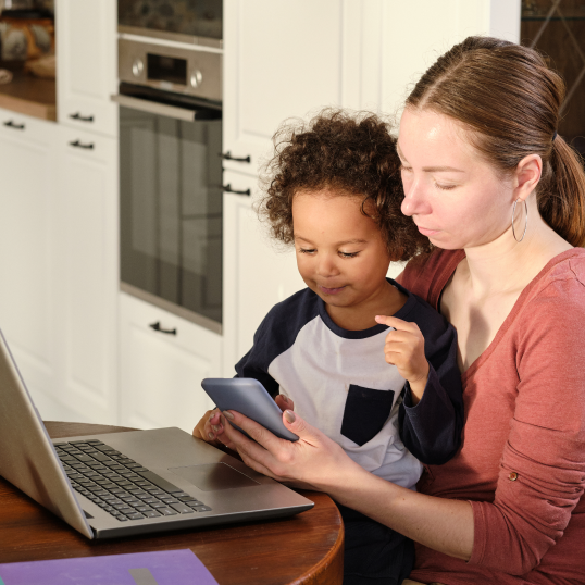 A woman and child are looking at a cell phone in front of a laptop