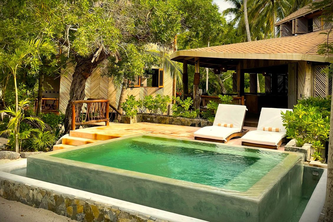 Piscina en entorno tropical con tumbonas y un pequeño edificio.