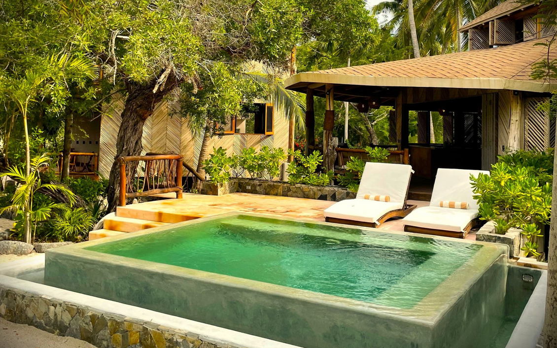 Piscina en entorno tropical con tumbonas y un pequeño edificio.