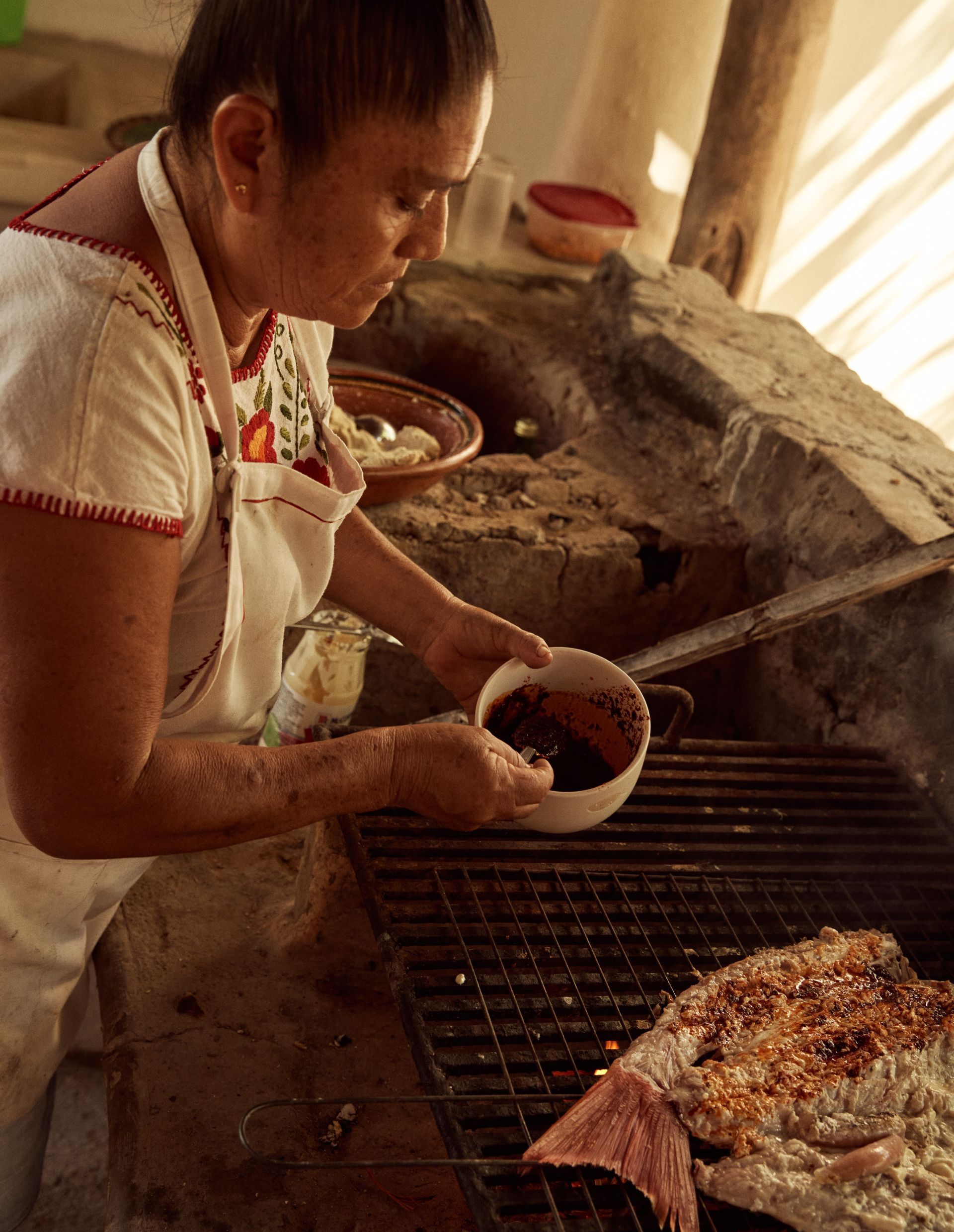 Mujer asando comida al aire libre, aplicando salsa. Luz solar, horno de piedra, delantal blanco.