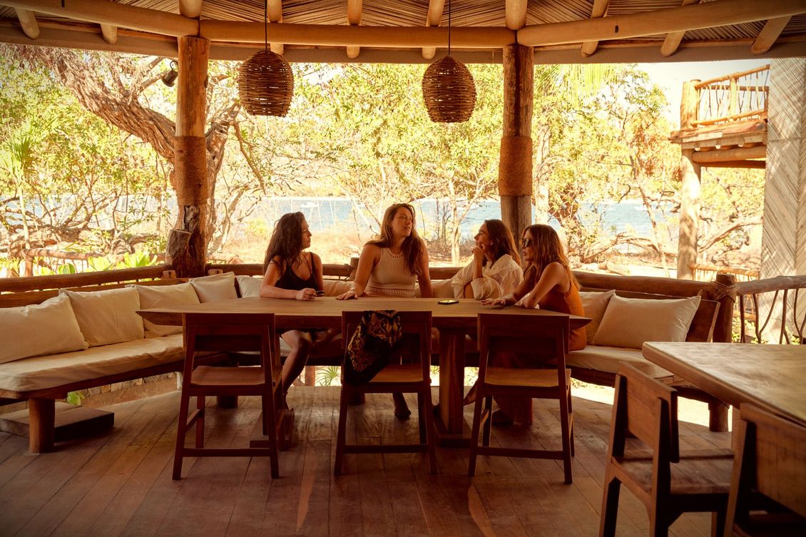 Cuatro mujeres sentadas alrededor de una mesa de madera en una terraza. Ropa beige claro. Entorno natural.