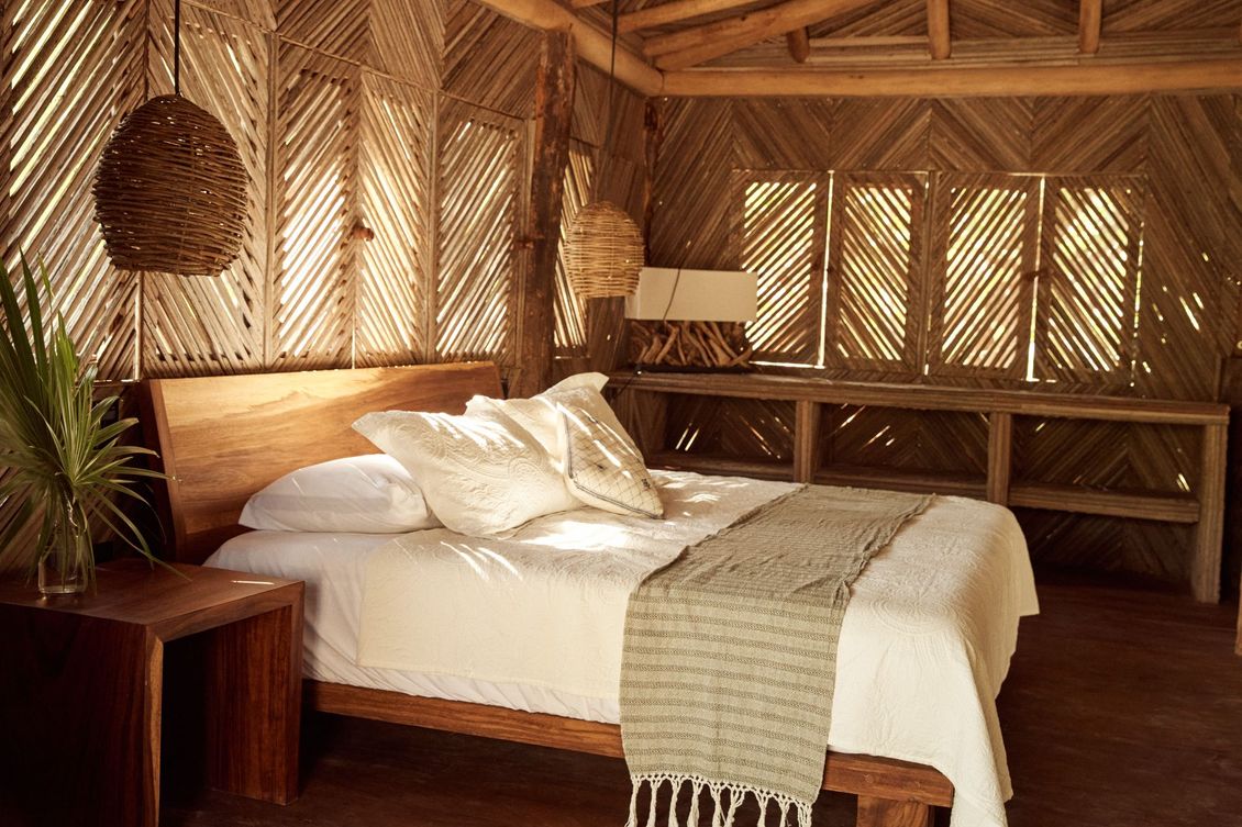 Dormitorio con cama de madera, paredes tejidas y lámparas colgantes. La luz del sol entra iluminando la cama.