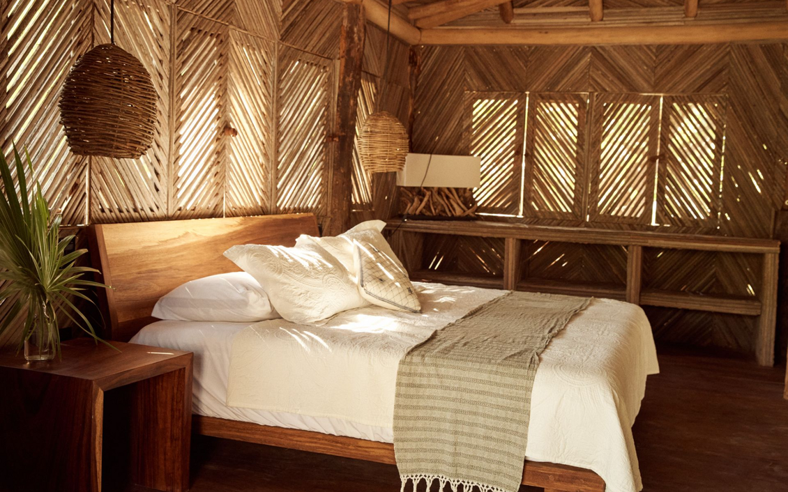 Dormitorio con cama de madera, paredes tejidas y lámparas colgantes. La luz del sol entra iluminando la cama.