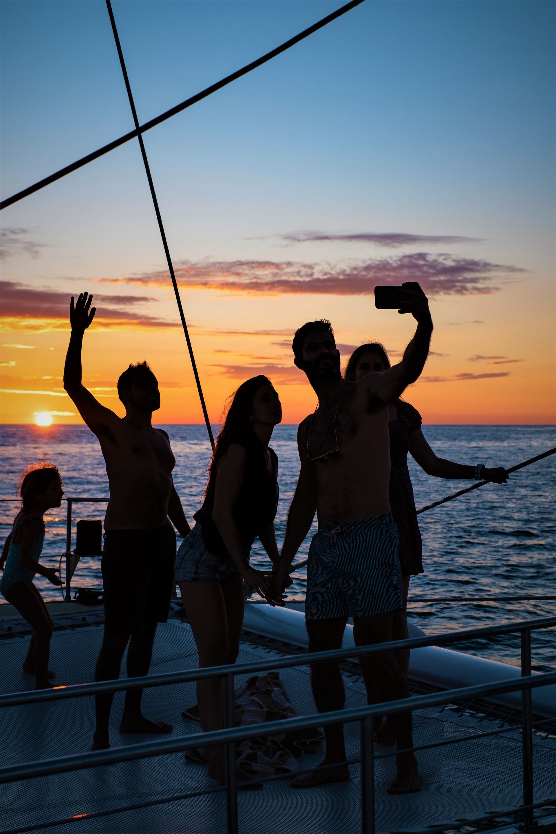 Grupo de siluetas tomándose un selfie en un barco al atardecer, con olas.