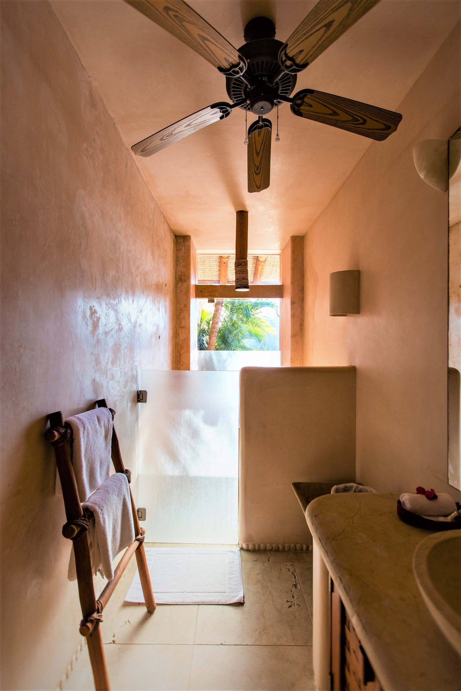 Baño con paredes beige, vigas de madera y ventilador de techo. La luz del sol entra a raudales por una abertura.